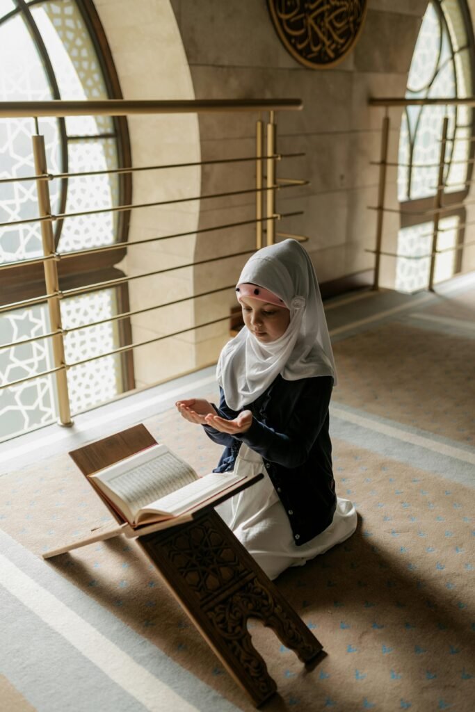 A young Muslim girl in a hijab prays indoors beside an open Quran on a stand.